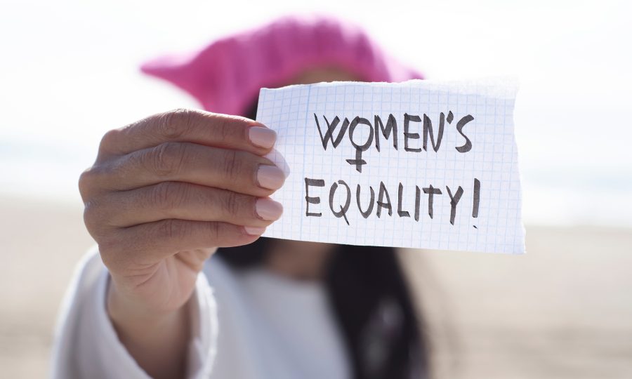 closeup of a young caucasian woman outdoors wearing a pink pussycat hat showing a piece of paper in front of her face with the text womens equality written in it